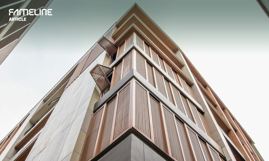 This image showcases the exterior of a modern multi-story building, featuring a dynamic façade with moveable sun shading solutions. The architecture combines sleek, clean lines with the warmth of wooden slats, which are part of a sun control system, possibly indicating a focus on energy-efficient building design. The angle of the photograph, looking up towards the sky, emphasizes the building's height and the thoughtful integration of these shading elements, suggesting a blend of functionality and aesthetic appeal in urban design. The visible branding "FAMELINE" suggest that this image might be used to promote an article or discussion about the benefits of smart architectural design in energy efficiency.