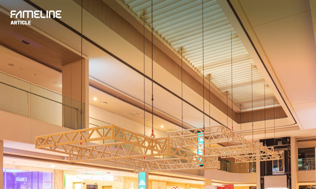 In the image, we see an expansive indoor space, likely part of a commercial or public building, featuring high ceilings and a sophisticated architectural design. The focal point is an innovative sunshade system, suspended elegantly above the space, complementing the wooden accents on the ceiling and the modern lighting setup. The sunshades are meticulously aligned, demonstrating both functionality and modern aesthetics, contributing to a well-lit, open, and airy atmosphere. This setting could be a lobby, an atrium, or a part of a shopping mall, where such architectural details not only enhance the beauty of the space but also improve the environmental comfort for visitors.