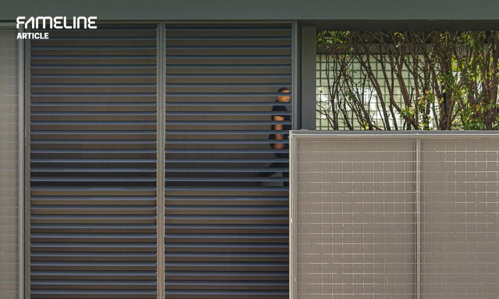 The image showcases an outdoor setting with a modern moveable louver system installed on a building facade. This louver system, possibly from the FAMELINE brand, is designed to offer flexible control over light and air flow, contributing to the building’s energy efficiency. The louvers are in a closed position, providing shade and privacy while still allowing glimpses of the greenery outside. The gray tones of the louvers match well with the building’s modern aesthetic, while the tiled wall in the foreground adds texture to the scene. This solution exemplifies contemporary architectural design that marries form with function, enhancing the building’s environmental performance and user comfort.
