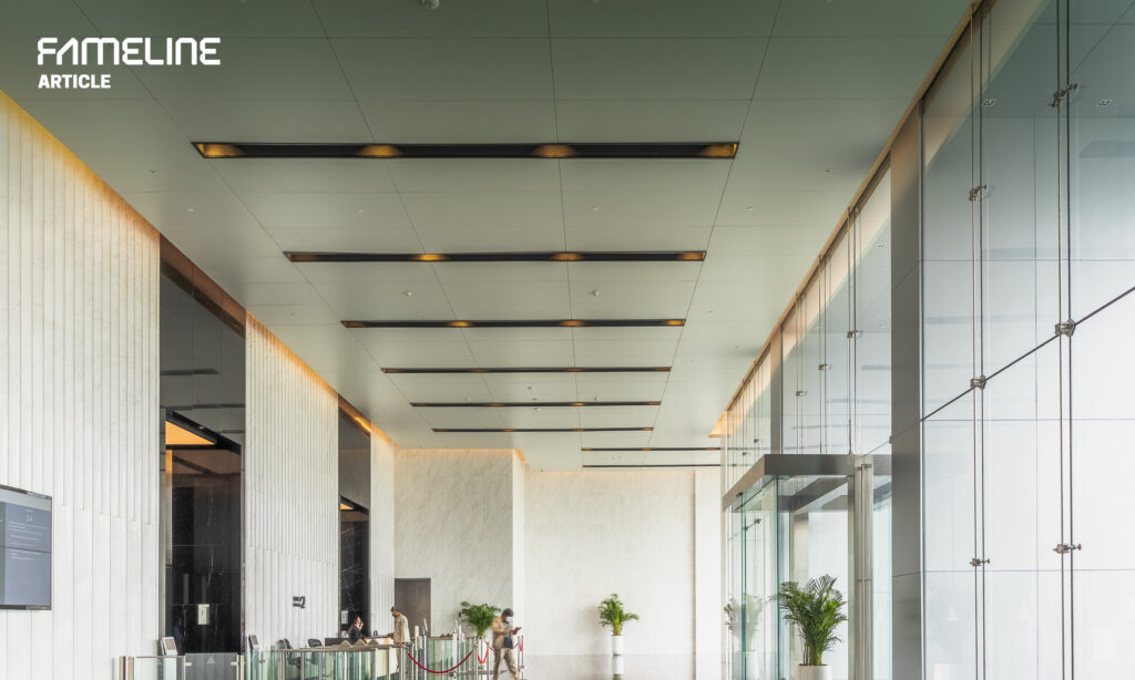 The photo showcases a sleek and modern interior hallway within a commercial or office setting. The hallway is characterized by a long perspective view framed by tall glass panels on one side, leading the eye towards the background. Overhead, the 'Mega+ Ceiling' system by FAMELINE is featured, with its neat rows of linear lighting integrated into the ceiling panels providing a warm, ambient glow. The clean architectural lines, the ample use of glass, and the minimalist lighting design contribute to an atmosphere of contemporary elegance and efficiency. This design choice underscores FAMELINE's commitment to combining aesthetic appeal with practicality, evident in the careful balance of natural and artificial lighting to create an inviting and productive environment.