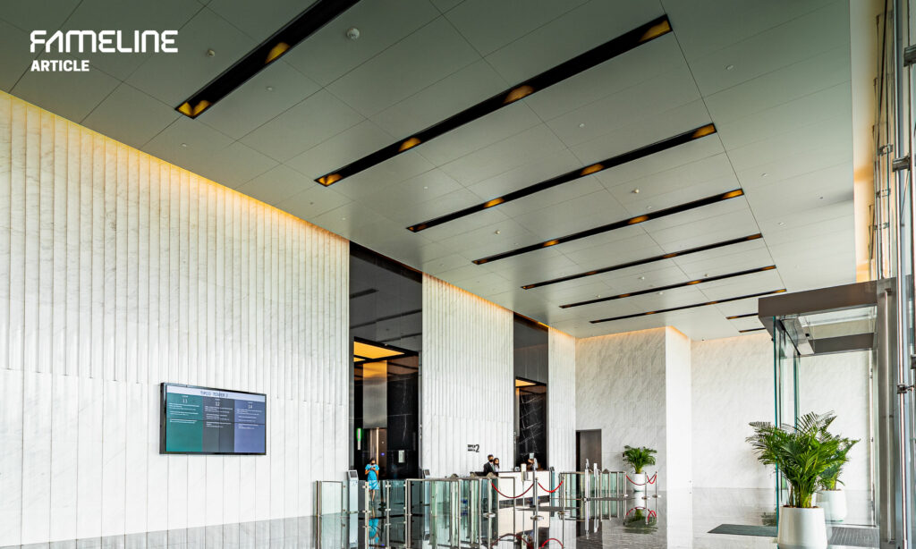 This image features a modern and sophisticated interior space, possibly a lobby or an entrance area of a contemporary building. The design integrates FAMELINE's 'Mega+ Ceiling' panels, which are aligned with precision, enhancing the architectural detail and adding a touch of modernity. The ceiling panels are interspersed with soft, warm lighting, which complements the natural light streaming in through the glass entrance. The interior is defined by clean lines and a neutral color palette, with marble walls that add a luxurious feel to the space. The open design, coupled with strategic lighting, creates a welcoming atmosphere that is both functional and aesthetically pleasing. Overall, this setting exemplifies FAMELINE's focus on combining functionality with high-end design to create elegant, energy-efficient spaces.