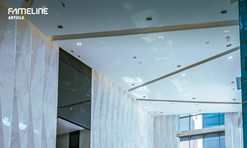 This image features a modern building's interior showcasing an expansive space with a high ceiling. The ceiling is adorned with strategically placed lighting fixtures that enhance the bright and airy ambiance. Marble walls with distinctive veining patterns line the sides, emphasizing opulence and grandeur. The use of glass walls introduces natural light and a sense of openness, while sleek, dark accents provide a contemporary contrast. The overall design conveys a blend of luxury and modernism, with attention to detail that speaks to FAMELINE's dedication to aesthetic appeal and quality in architectural design.