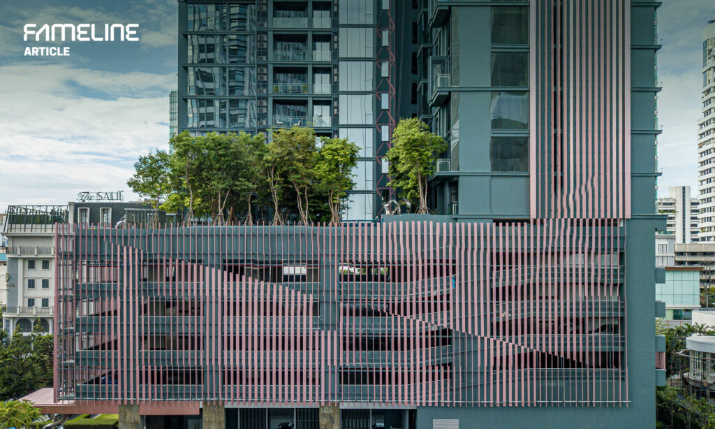 This image features a modern building facade adorned with pink vertical louvers, integrating both aesthetic and functional design elements. The louvers provide sun shading and enhanced privacy while allowing for natural ventilation. Behind the louvers, lush greenery on terraces adds a touch of nature, contrasting with the urban setting visible in the background, including a mix of high-rise buildings and a sign reading "The Sail." This integration of green spaces on urban buildings highlights a sustainable approach to modern architecture.