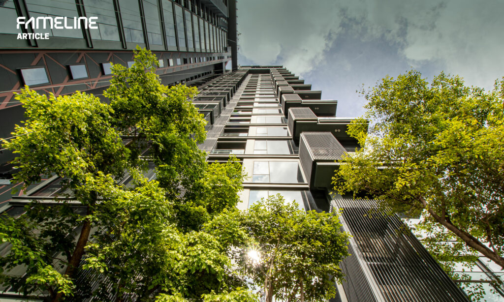 This image captures the modern architecture of a high-rise building accentuated with sun and ventilation louvers. The design integrates green, lush trees that seem to merge seamlessly with the urban structure, enhancing its aesthetic and environmental quality. The perspective is from ground level looking up towards the sky, emphasizing the building's impressive height and the sustainable design elements that contribute to energy efficiency and occupant comfort. This image highlights the practical application of environmental sustainability principles in contemporary urban development.