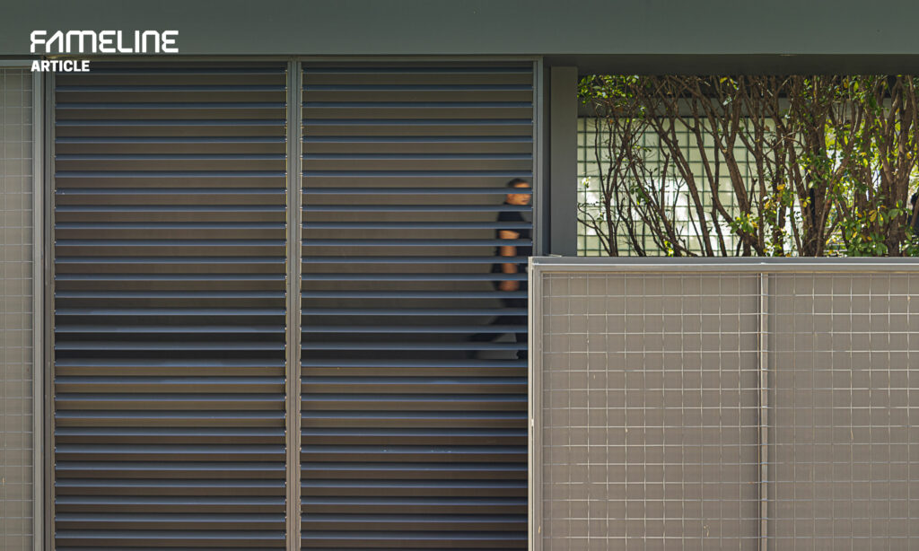 This image showcases a section of a building facade with dark gray louvers, which are part of a sun shading system designed to enhance both privacy and ventilation. A person is partially visible through the louvers, walking by, hinting at the human scale and functional use of the space. The background features a green, leafy area behind a glass barrier, adding a natural element to the architectural design. The overall setting is a blend of modern design and functionality, emphasizing the integration of sustainable building practices in urban environments.