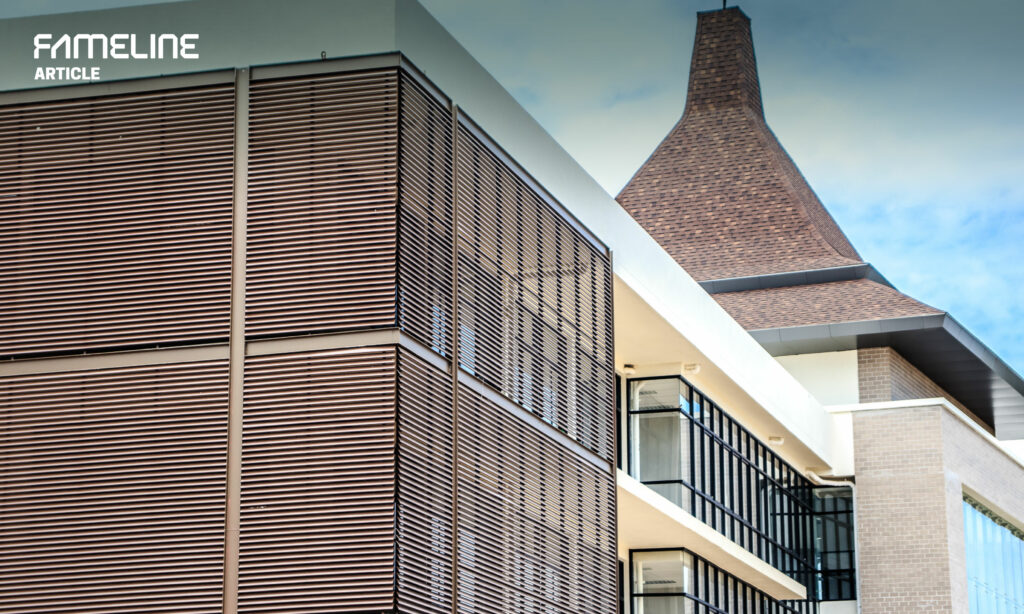 This image displays a section of a modern architectural structure featuring sun and ventilation louvers. The louvers are in a rich brown hue, contrasting with the building’s light-colored brick and white exterior. A distinctive, large brick chimney extends into the sky, adding a unique element to the building’s profile. The combination of functional and aesthetic design elements highlights the building's modern approach to blending traditional materials like brick with contemporary features for energy efficiency and climate control.